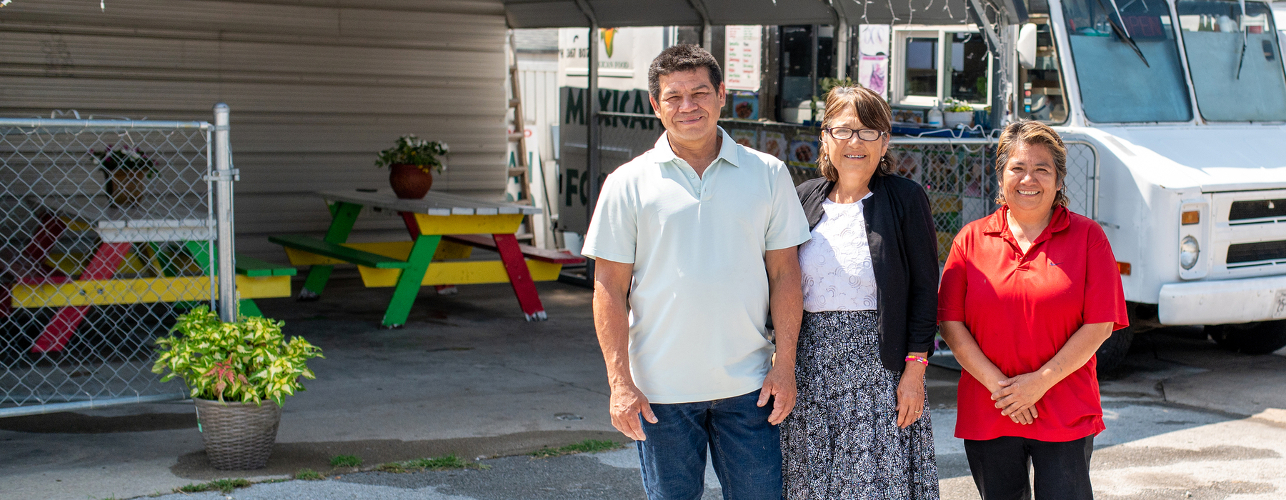 One Latino male and two Latina women stand outside a white food truck.