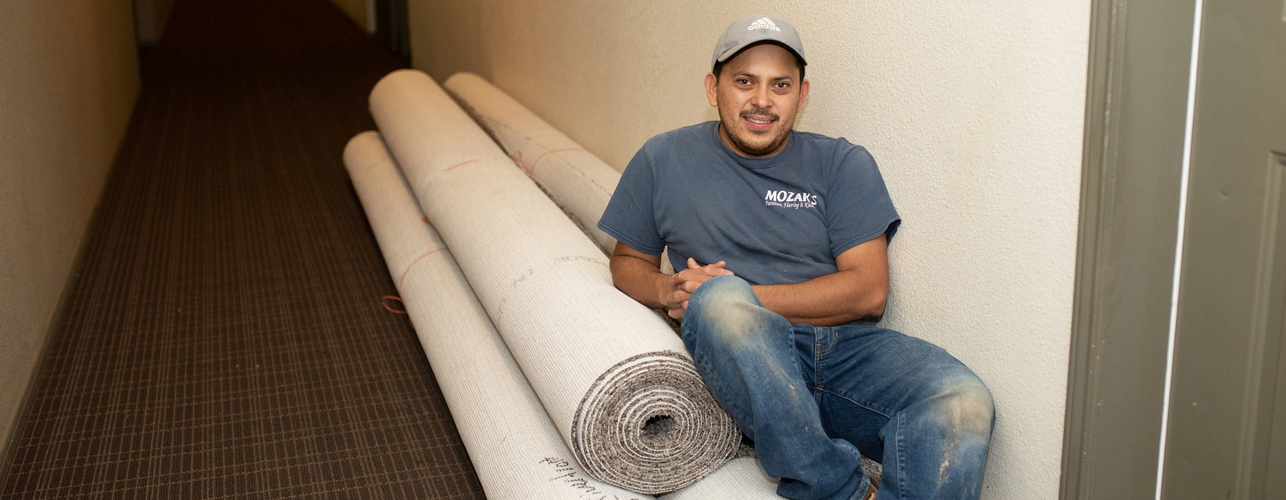 Hispanic male in grey shirt leans on a few rolls of carpet in a hallway