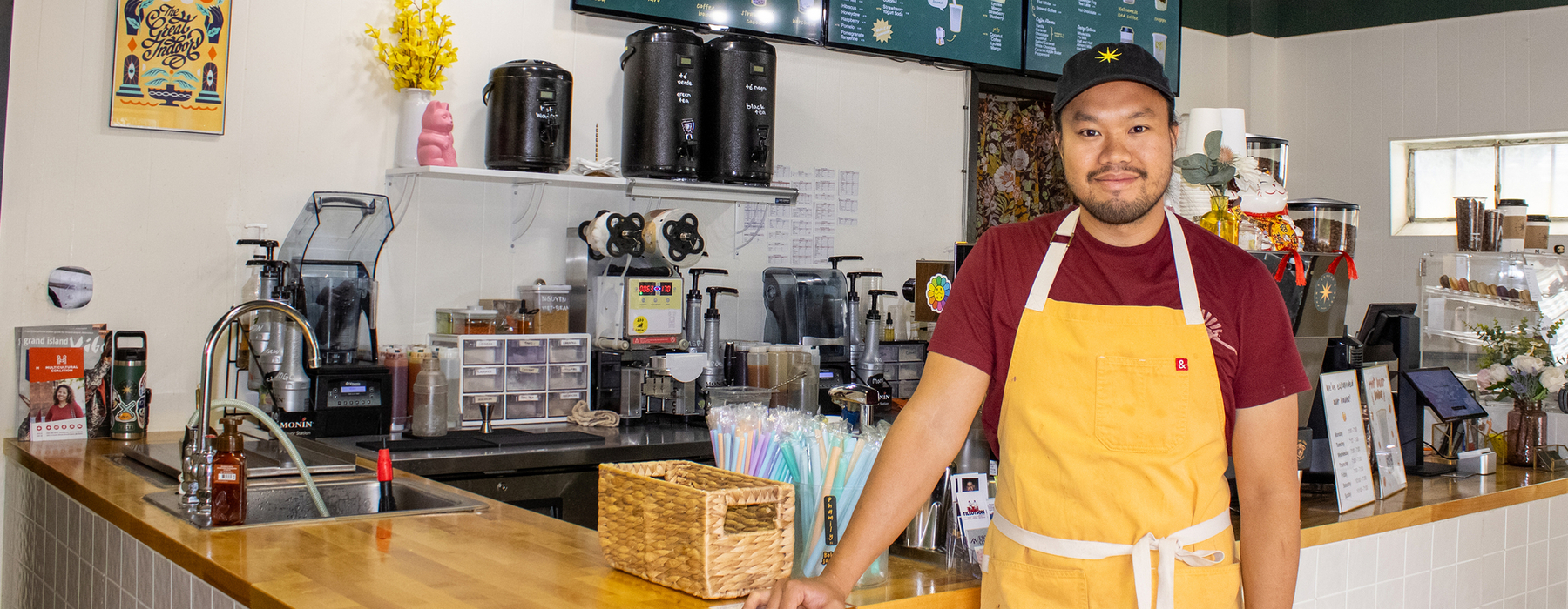 Male with black hat, red shirt, and yellow apron stands near a coffee shop stand indoors.