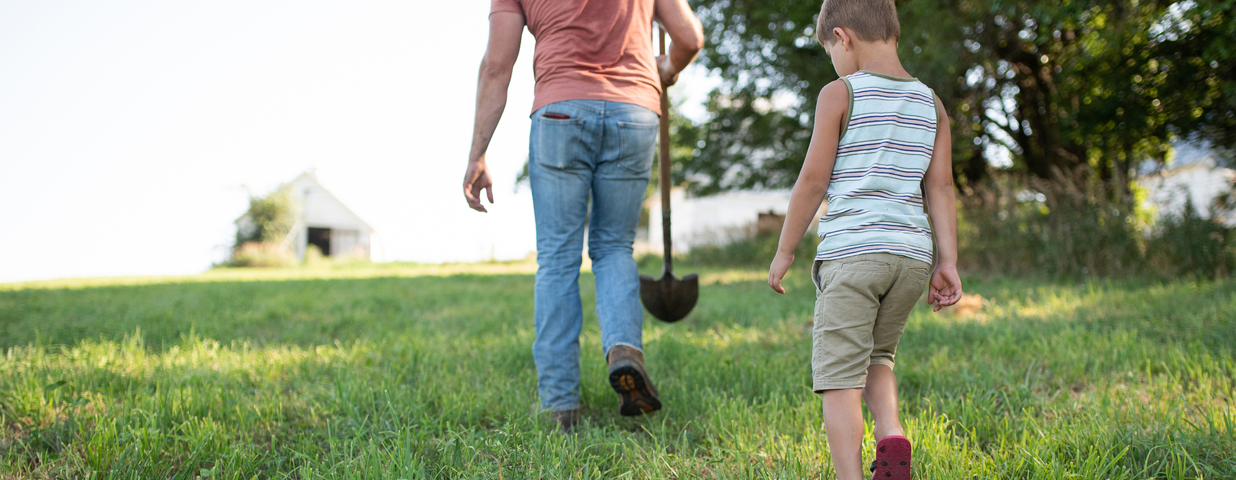 Un home amb texans, camisa de préssec, amb una pala camina per una pastura d'herba seguit d'un nen jove amb pantalons curts i una samarreta de tirants de ratlles blanques i marrons.
